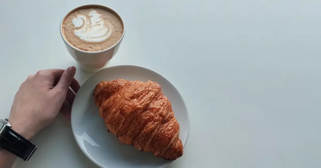 Coffee and a croissant on a white table next to a resting hand